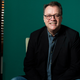 Russell T. Davies poses in a new photo released by the BBC to accompany the announcement of his return to Doctor Who in 2023. He's sitting, wearing a black suit jacket over a blue button down shirt and in front of a green paper background.
